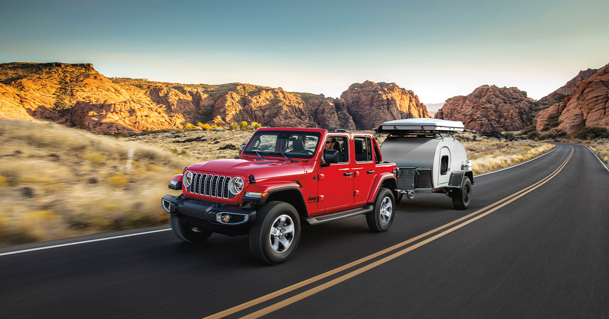 A red Jeep Wrangler towing a trailer on the road in a desert.