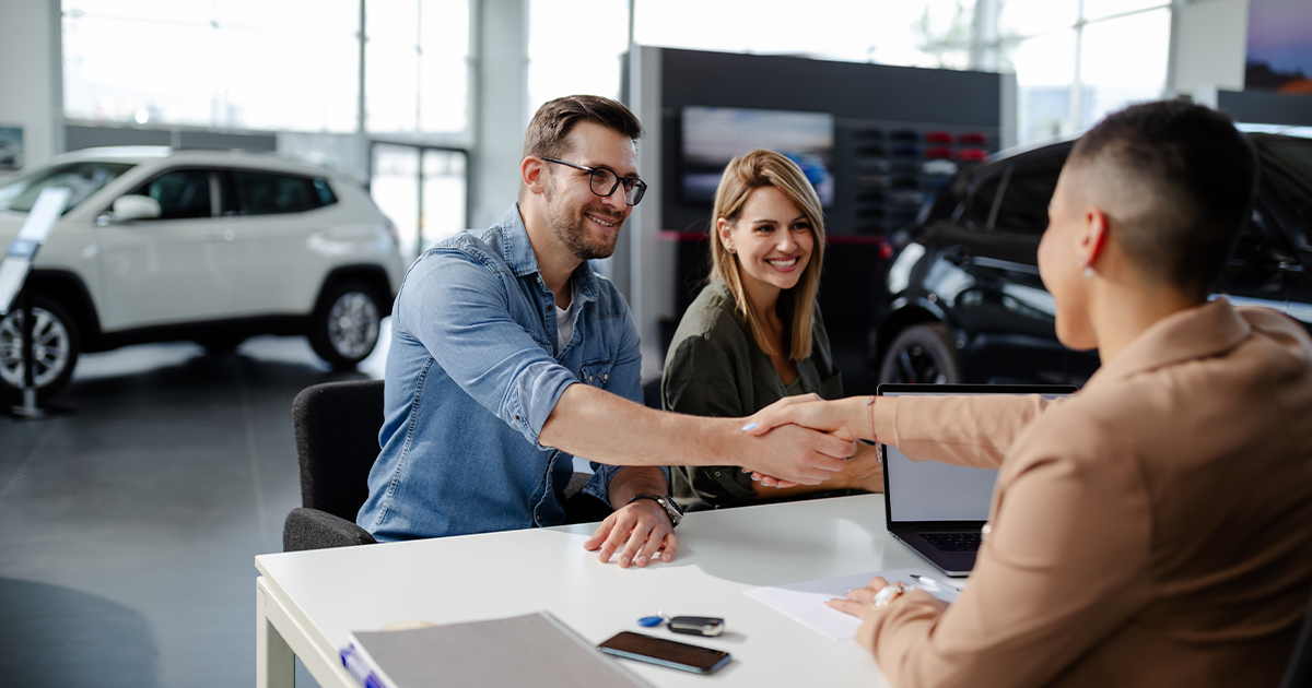 A couple shaking hands with a car salesperson at his desk in the car dealership showroom.